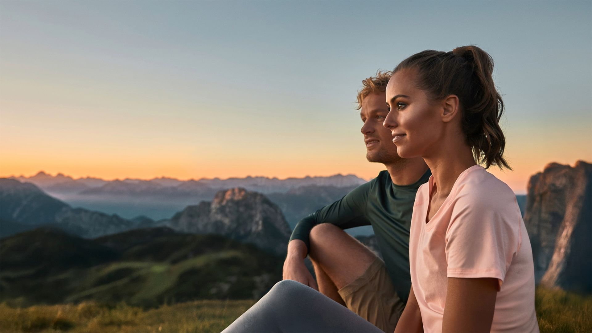 Paar sitzt auf Berggipfel und genießt den Sonnenuntergang über den Alpen.
