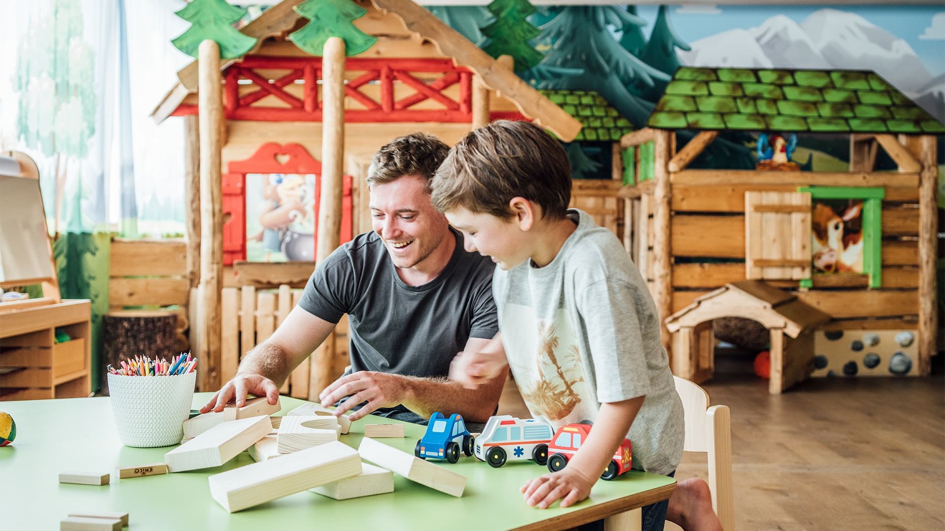 Vater und Sohn spielen gemeinsam mit Holzautos und -bausteinen an einem Tisch in einem Spielhaus.