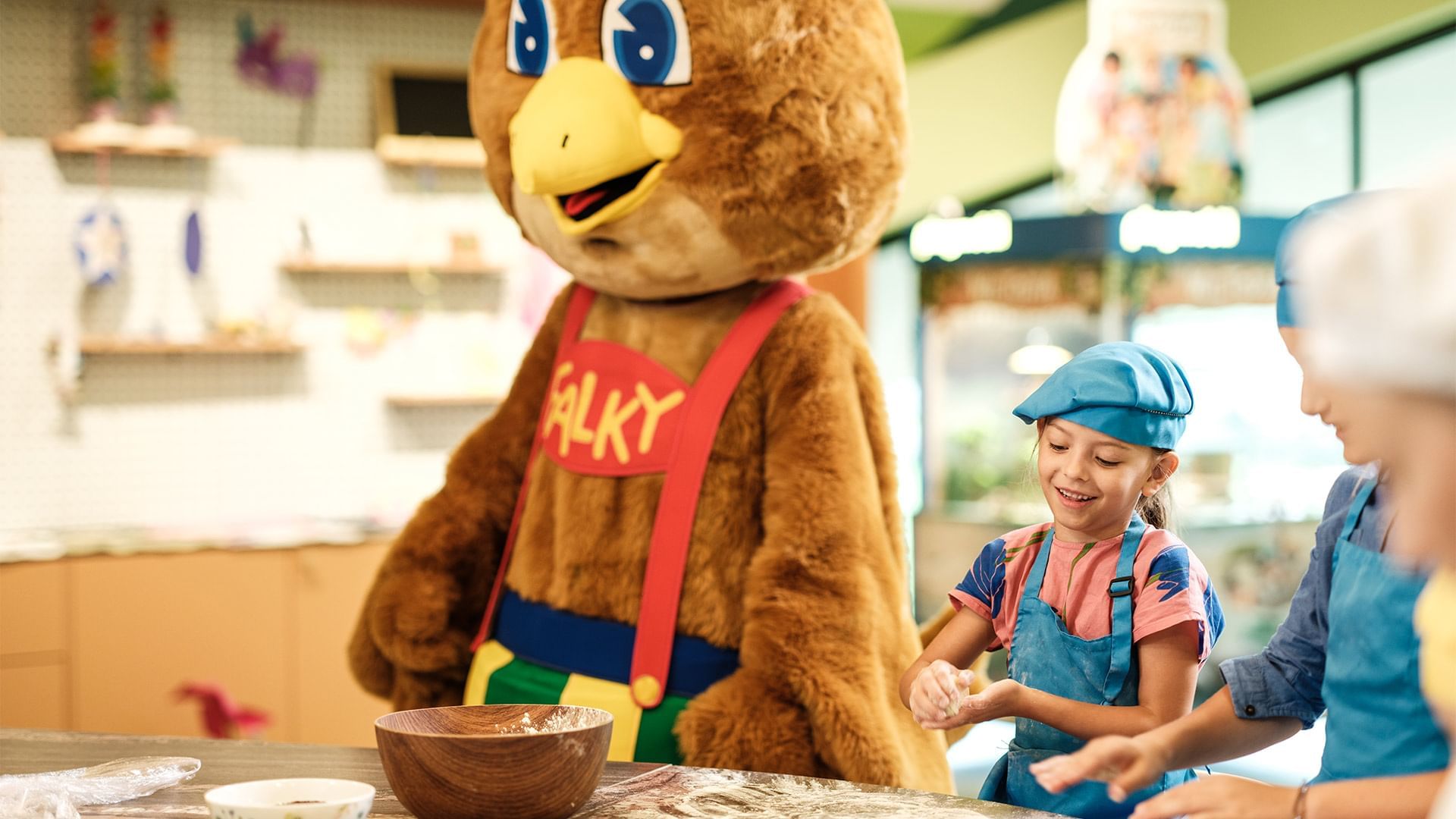 Ein Maskottchen mit dem Namen Falky steht neben zwei Kindern, die Teig kneten.