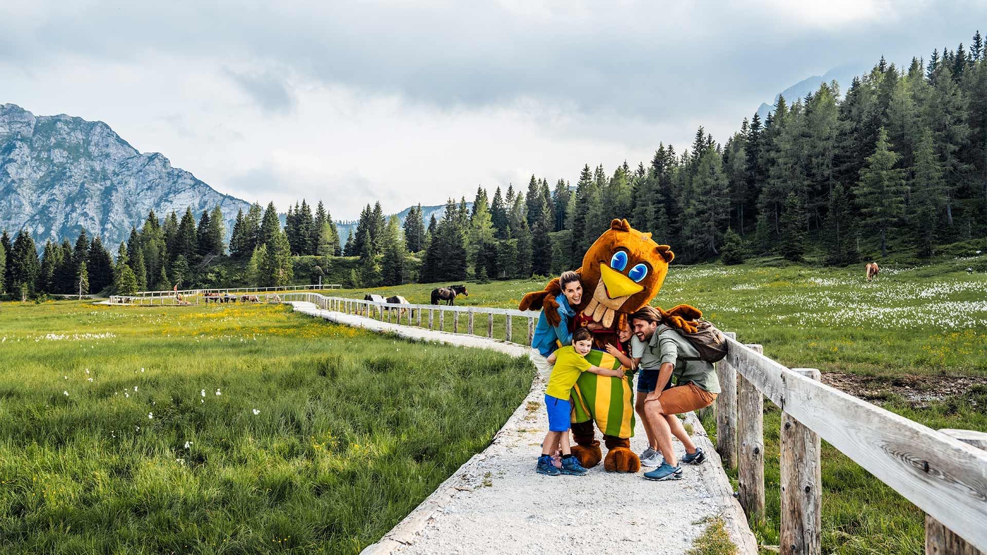 Familie und Maskottchen auf einem Pfad in den Alpen, umgeben von Wiesen und Wald.