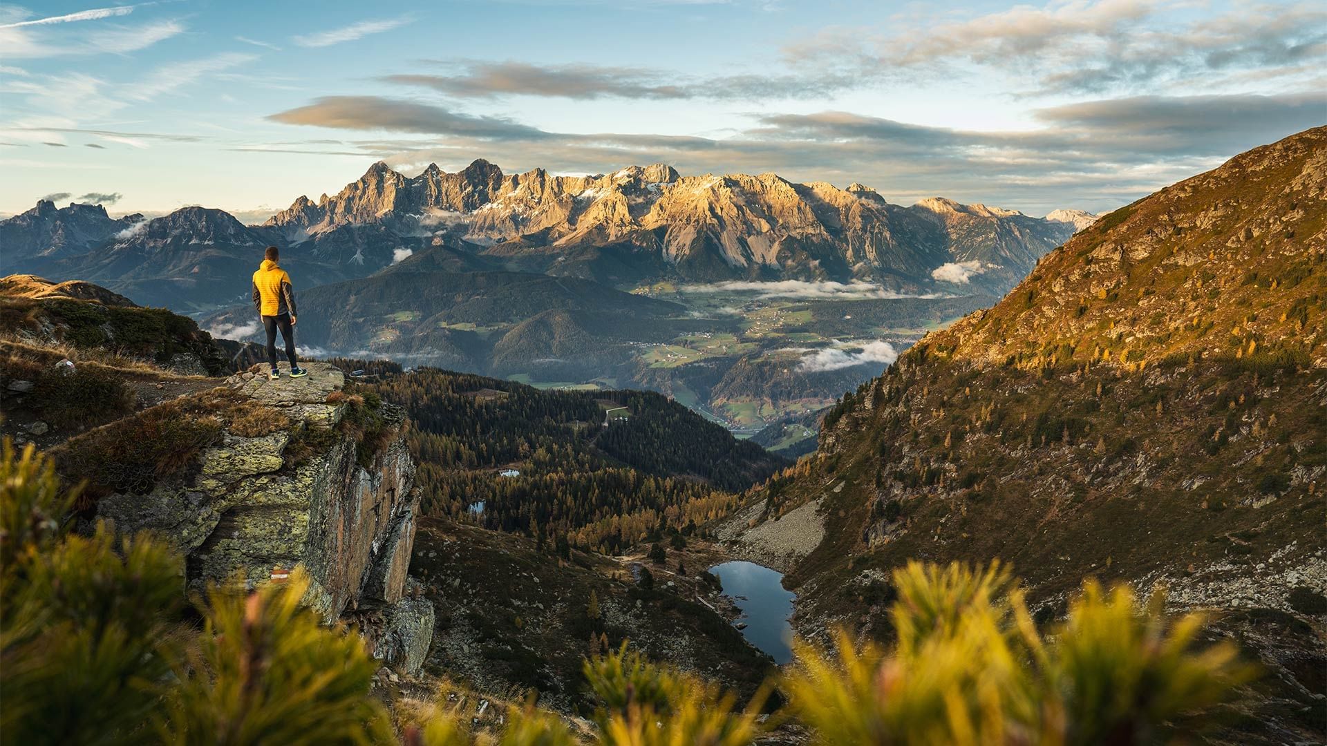 Mann in gelber Jacke auf Felsvorsprung mit Blick auf Bergkette und Tal in der Abendsonne.