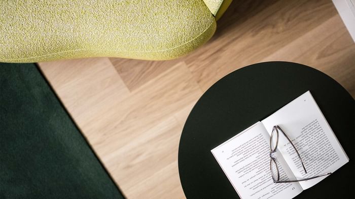 Schwarzer Tisch mit Buch und Brille, neben einem grünen Sessel auf Holzboden.