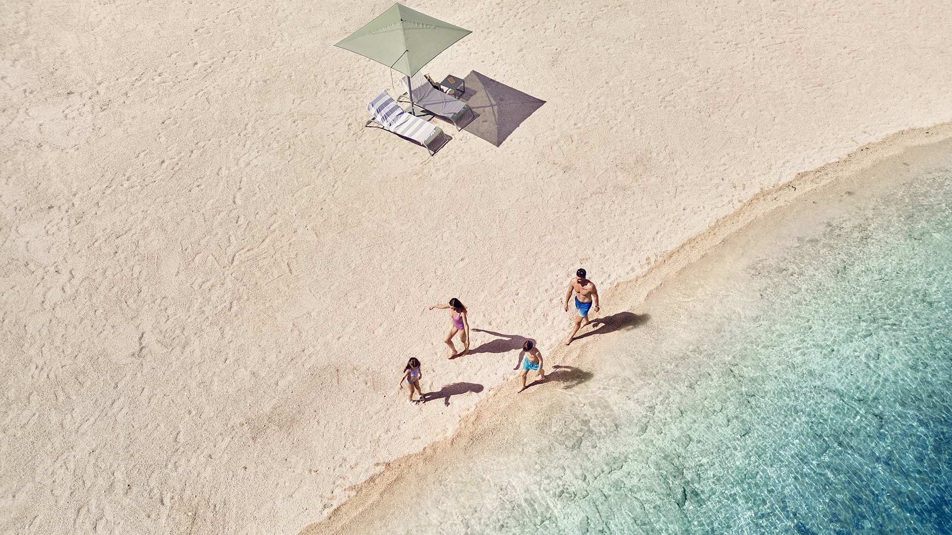 Eine Familie spaziert am Sandstrand, der an klares, blaues Wasser grenzt, mit Liegen und Sonnenschirm.