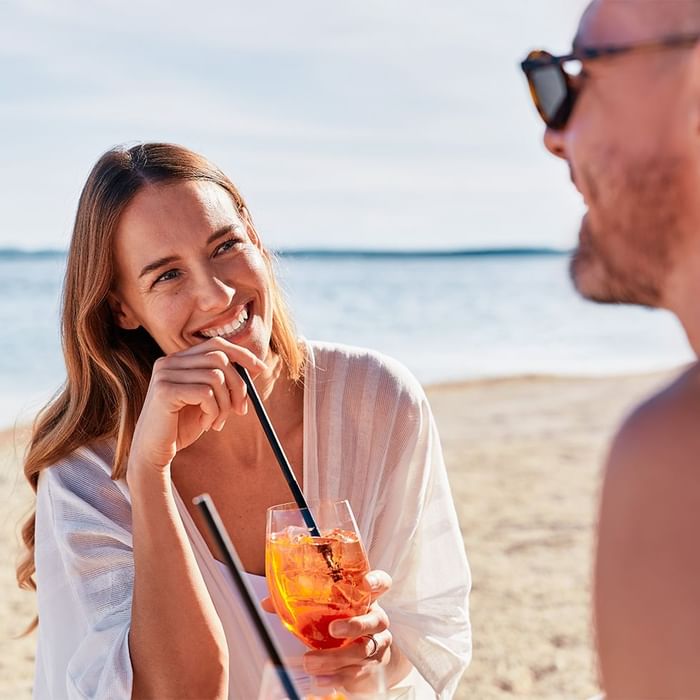 Frau mit Cocktail und Strohhalm lächelt am Strand neben einem Mann in Sonnenbrille.
