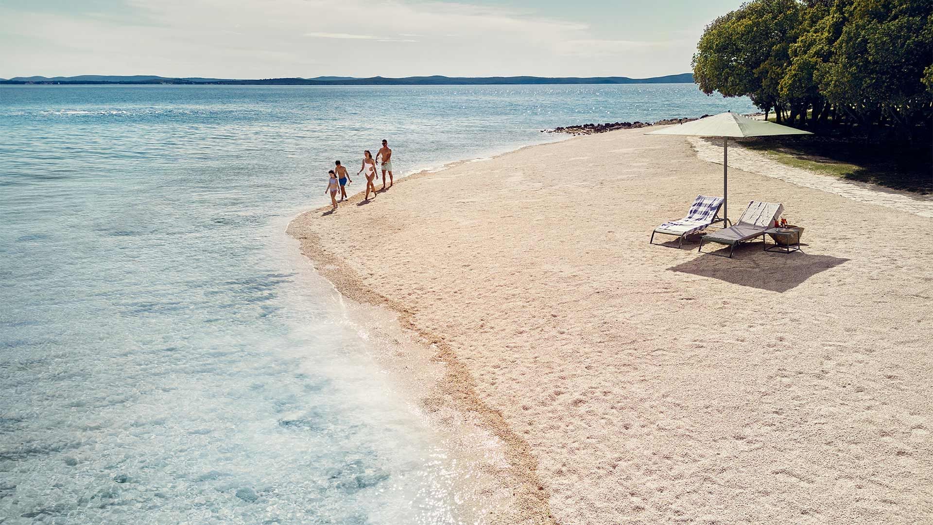 Strandszene mit Familie, Liegen, Sonnenschirm und kristallklarem Wasser unter einem wolkenlosen Himmel.