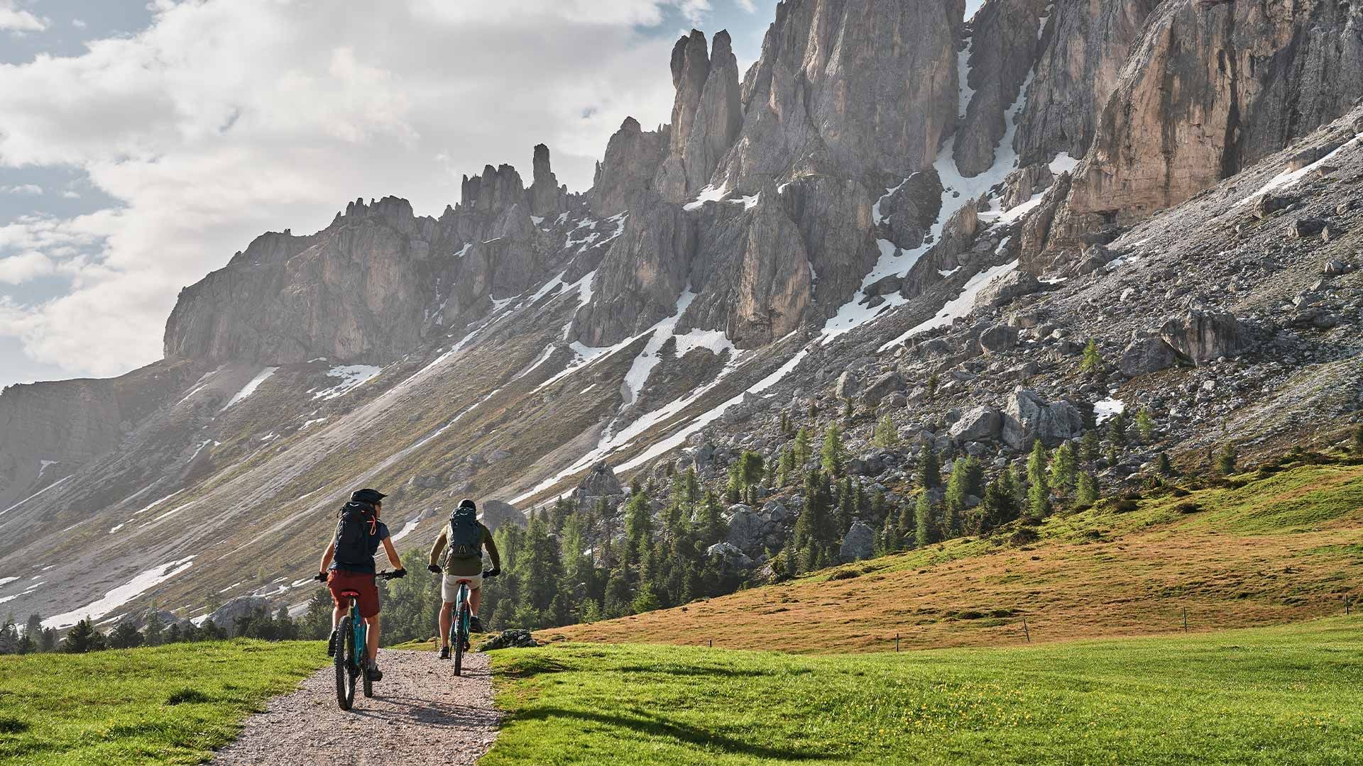 Zwei Radfahrer mit Rucksäcken auf einem Pfad vor einem Berg mit Schnee und Wolken.