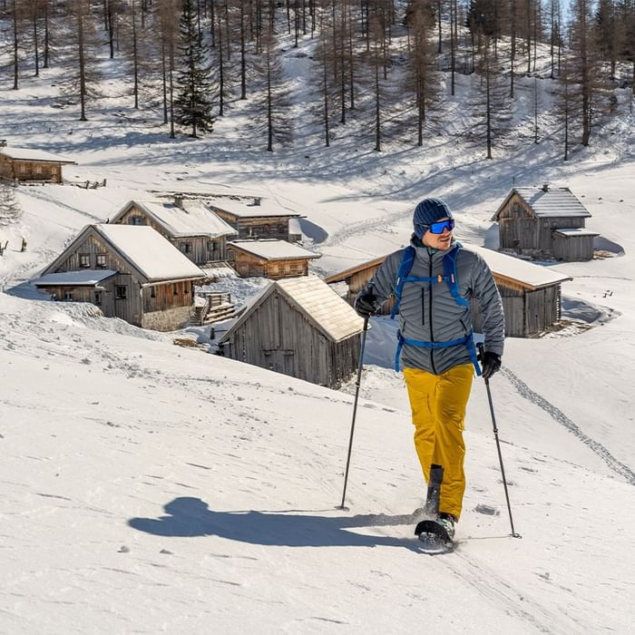 Snowboarder mit gelber Hose und grauer Jacke, der auf einem schneebedeckten Berg mit Holzhütten im Hintergrund steht.