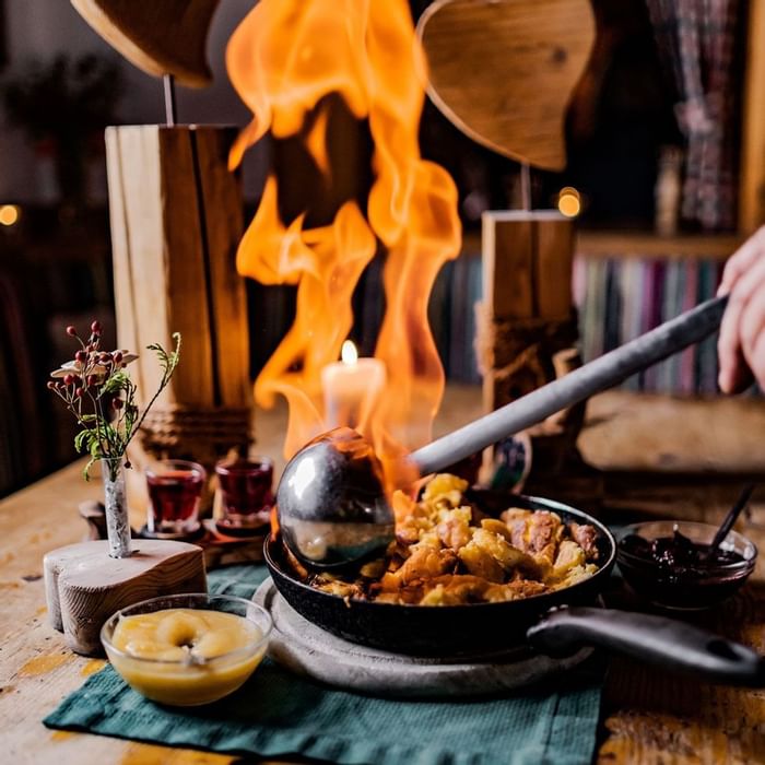 Flammendes Gericht mit Löffel in brennendem Topf, umgeben von Gläsern und Dekoration auf einem Tisch.
