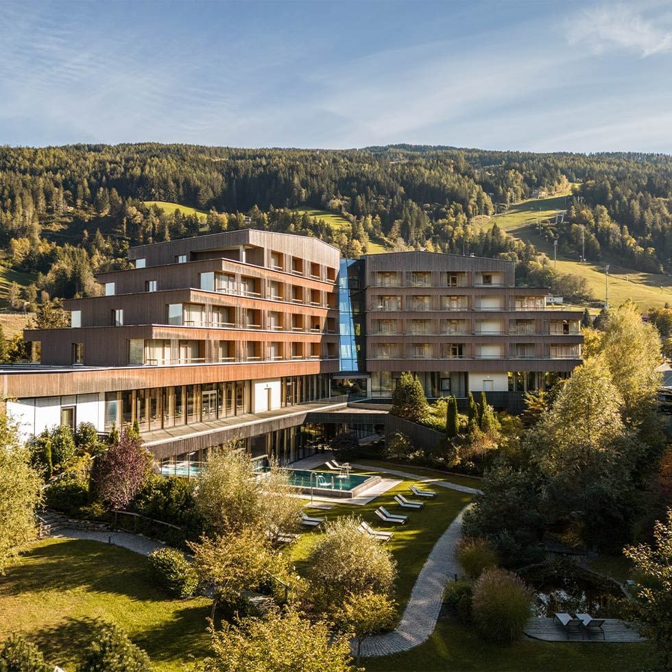 Modernes Hotel mit Pool und Liegen, umgeben von Bäumen und einem Berg im Hintergrund.