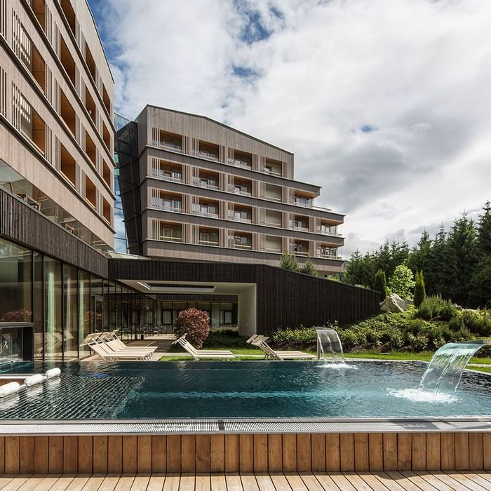 Modernes Hotelgebäude mit Außenpool, umgeben von Grün und Liegestühlen unter einem bewölkten Himmel.