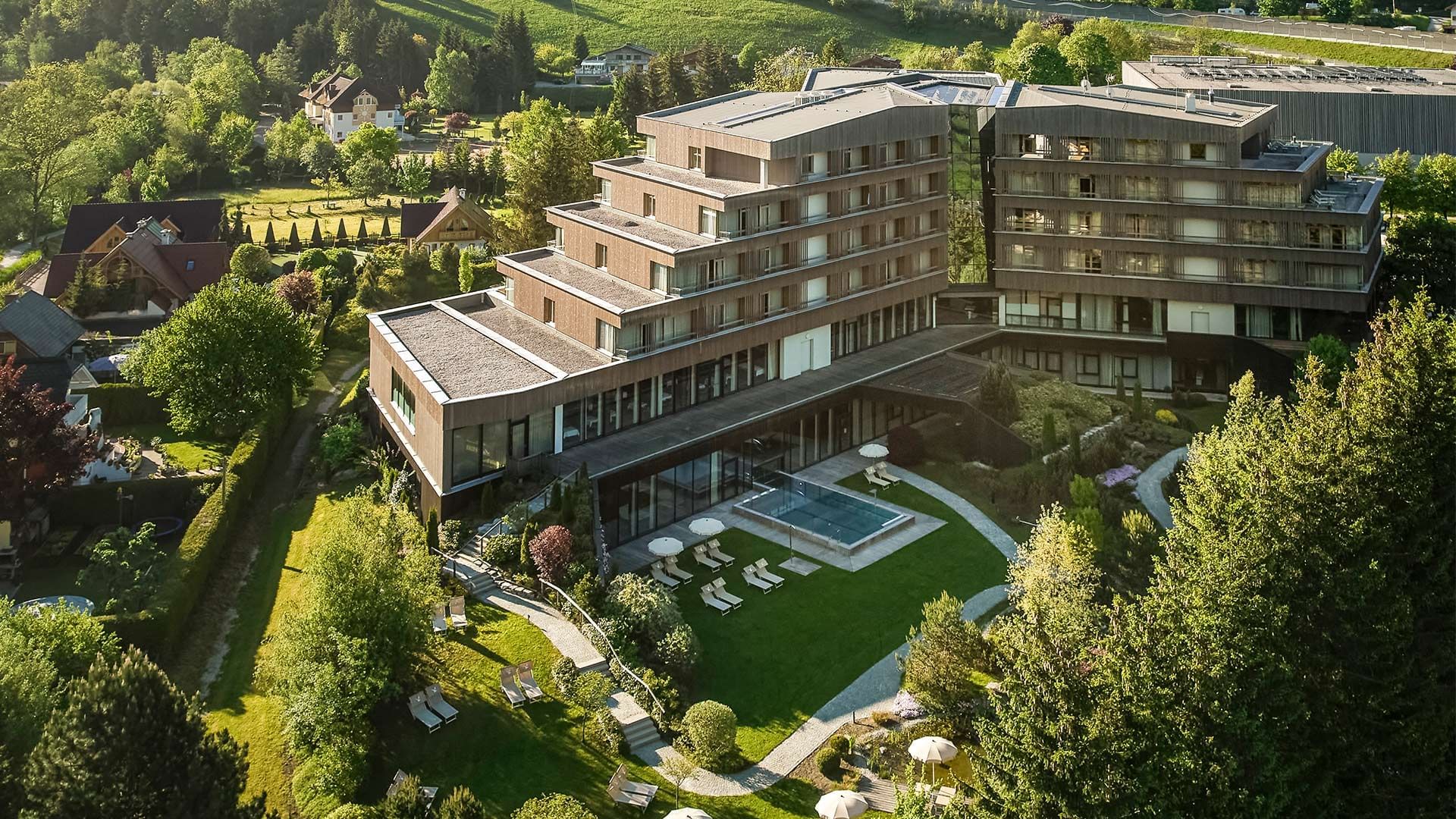 Vista aerea di un moderno hotel con piscina e giardino, circondato da alberi e case.