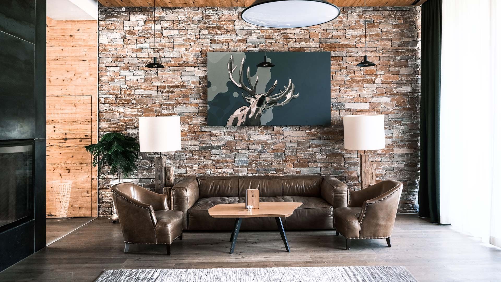 Wohnzimmer mit Steinwand, Hirschbild, Ledercouch, Couchtisch und zwei Sesseln neben Stehlampen.