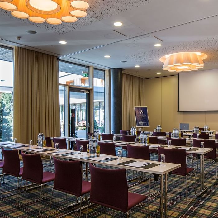 Konferenzraum mit Tischen, Stühlen, Leinwand, Präsentationstafel und Getränken auf den Tischen.