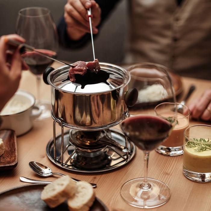 Personen, die Fleisch am Tisch mit Fonduetopf und Wein auf einem Holztisch essen.