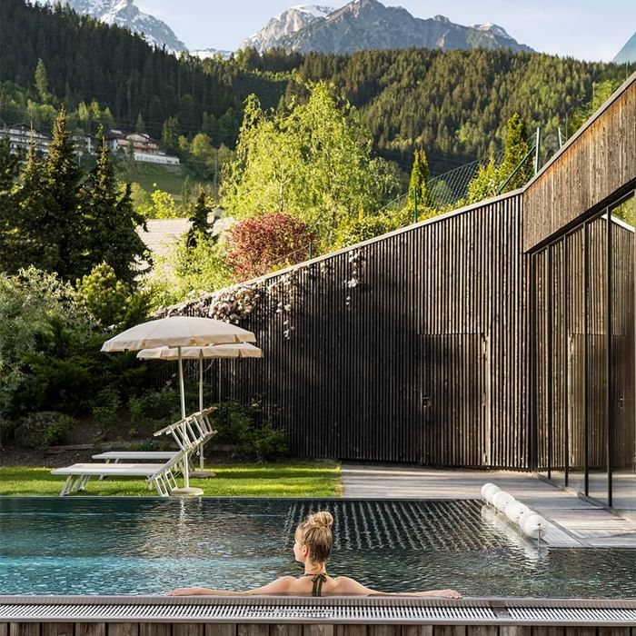 Frau entspannt sich im Schwimmbad mit Bergblick, umgeben von Holzbarrieren und Sonnenschirmen.