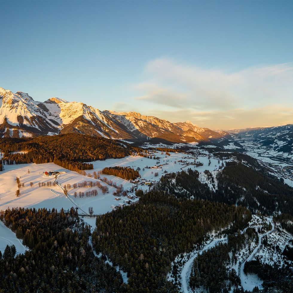 Luftaufnahme einer schneebedeckten Landschaft mit bewaldeten Hügeln und majestätischen Bergen im Hintergrund.