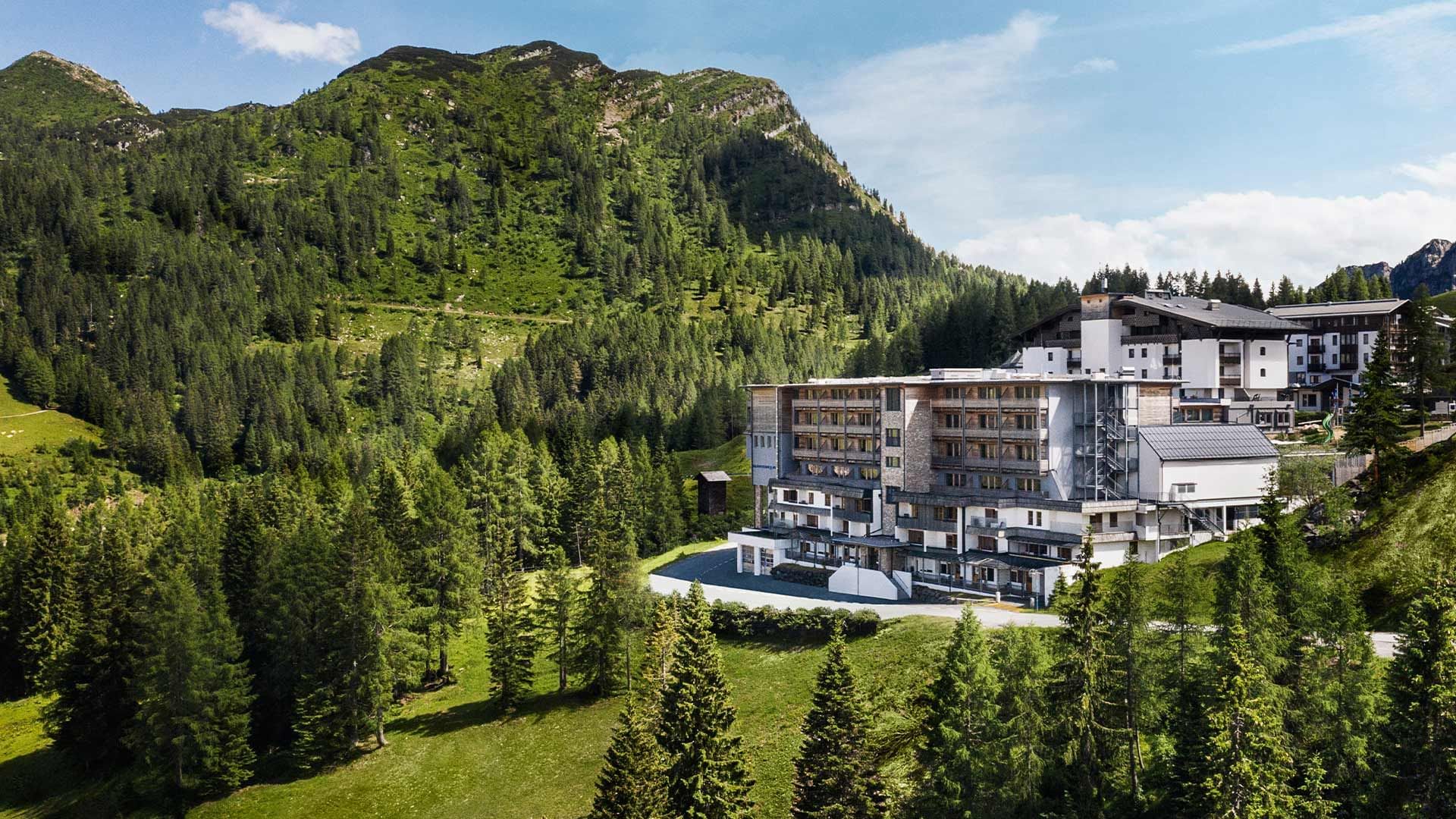 Modernes, mehrstöckiges Gebäude in den Alpen, umgeben von Bäumen und grünen Hängen.