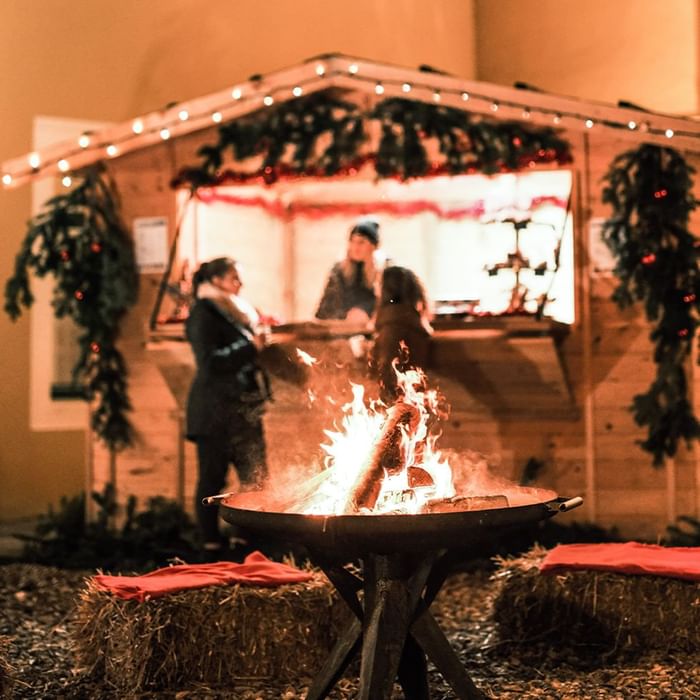 Menschen stehen vor einem Weihnachtsstand mit Glühwein und Feuerschale, umgeben von Strohballen und Lichterketten.