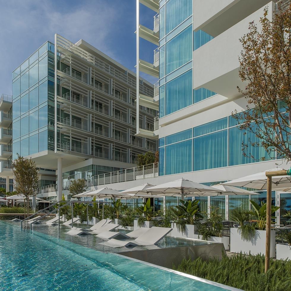 Modernes Hotelgebäude mit Pool, Liegen, Sonnenschirmen und Pflanzen unter blauem Himmel.