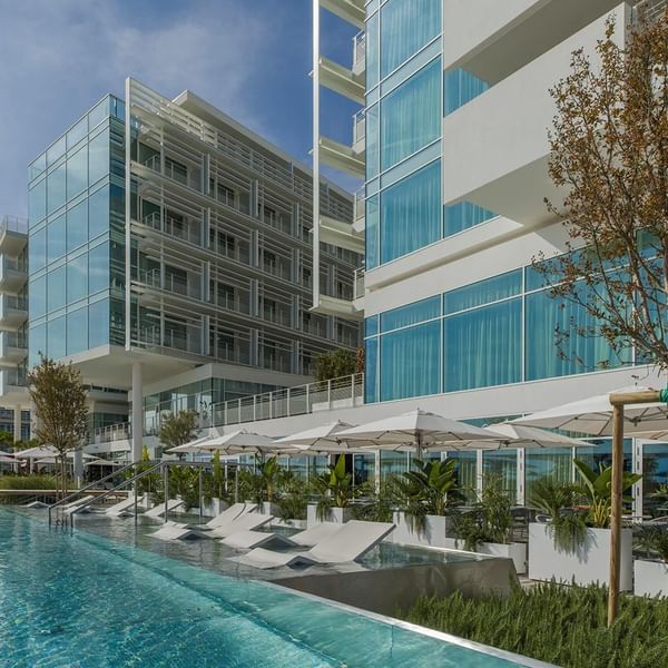 Modernes Hotelgebäude mit Pool, Liegen, Sonnenschirmen und Pflanzen unter blauem Himmel.
