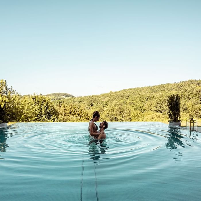Zwei Menschen schwimmen in einem Pool vor einer bewaldeten Hügellandschaft.