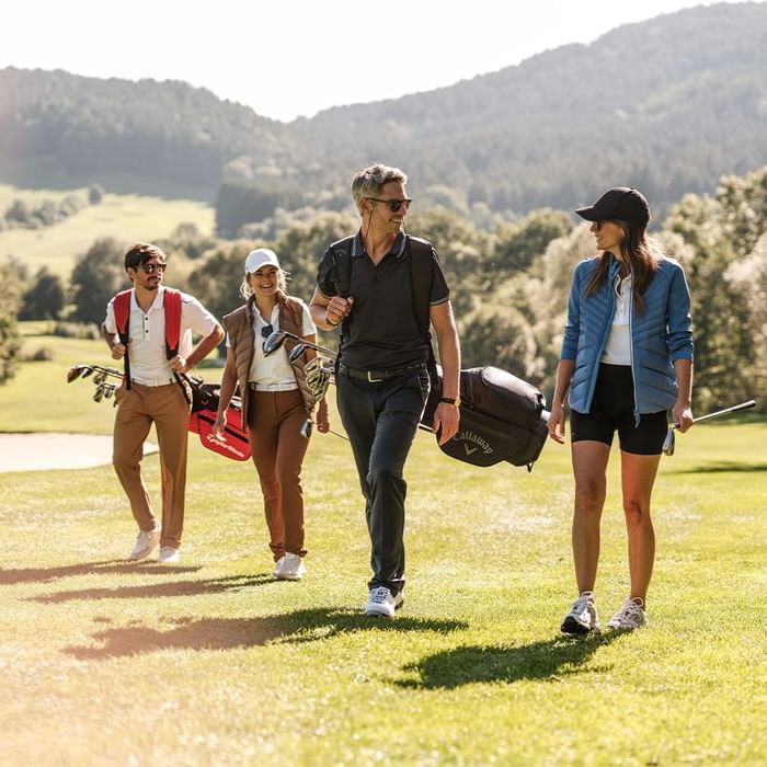 Vier Golfspieler mit Ausrüstung auf einem grünen Golfplatz vor Bergkulisse, alle tragen Sonnenbrillenille