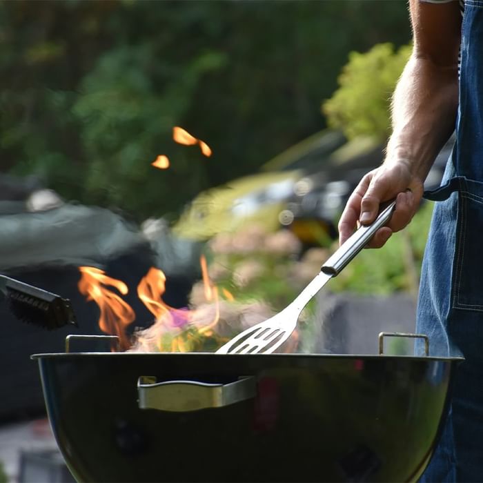 Mann in Jeansoveralls grillt mit einer Zange über einem brennenden schwarzen Grill im Freien.