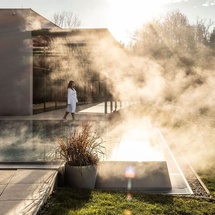 Donna in accappatoio bianco che cammina lungo un sentiero di vapore vicino a una piscina esterna.