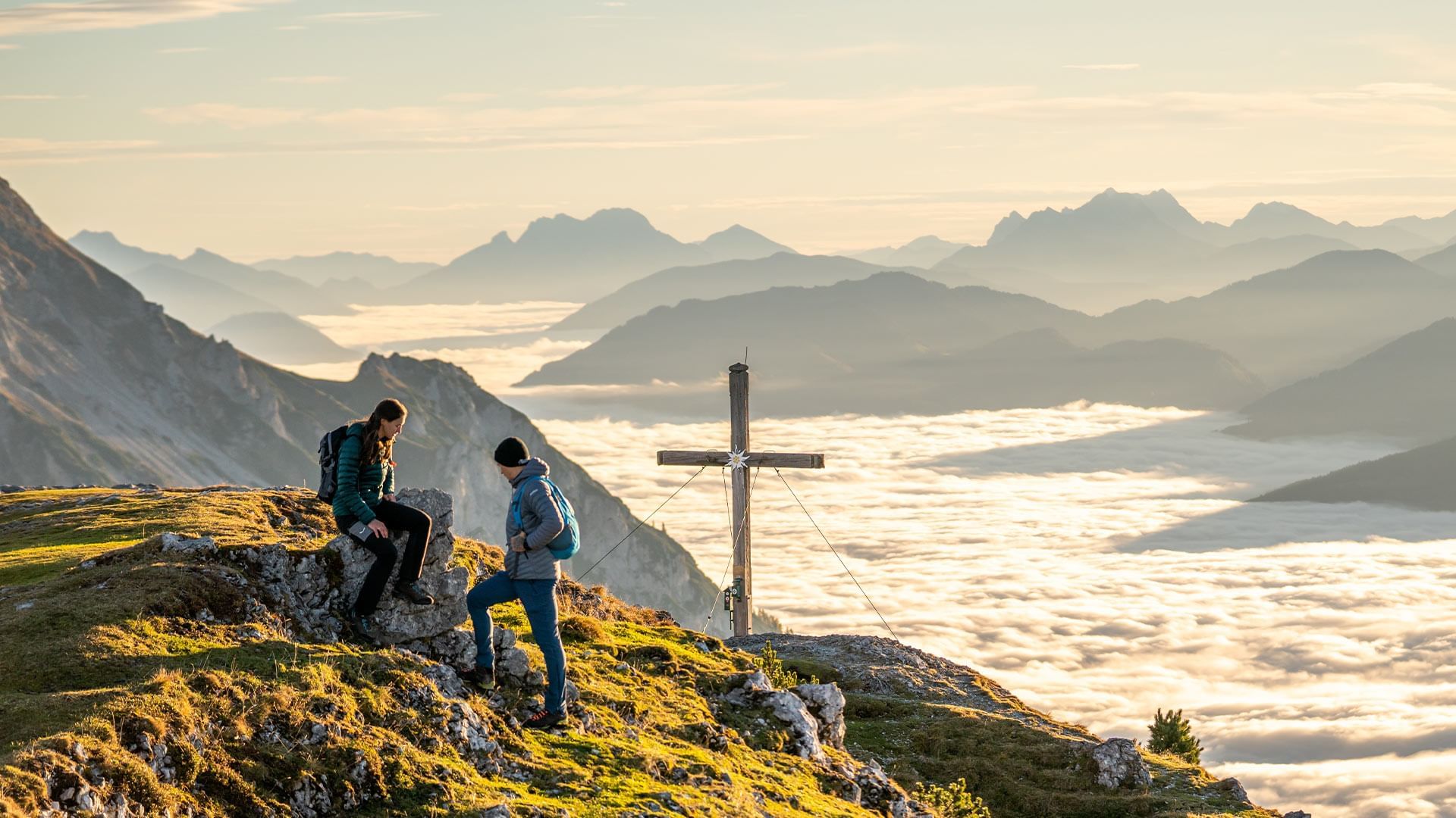 Zwei Wanderer auf Berggipfel mit Kreuz und nebligen Tälern im Hintergrund.