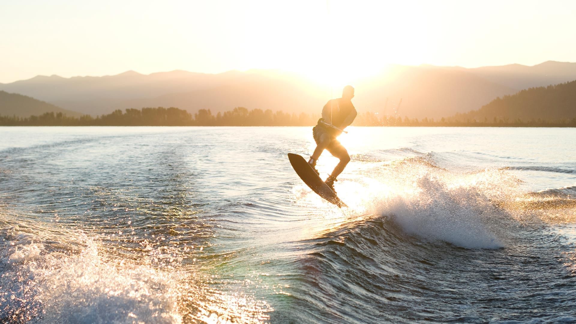 Mann auf Wakeboard über Wasserwelle, Sonnenuntergang über See mit Berglandschaft im Hintergrund.