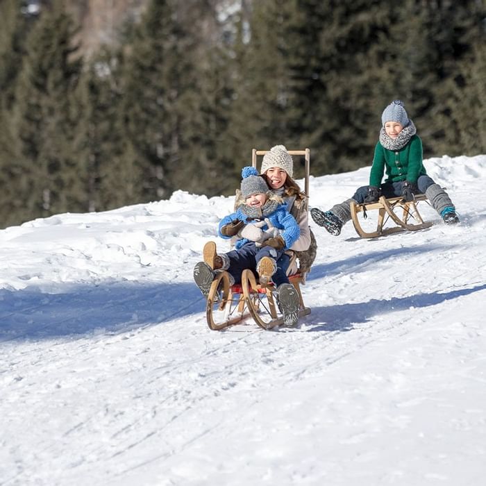 Mutter und Kind auf einem Schlitten, einem anderen Kind hinterherfahrend, auf einem verschneiten Hügel.