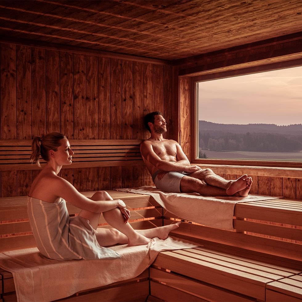 Zwei Personen entspannen in einer hölzernen Sauna mit Ausblick auf eine Landschaft.