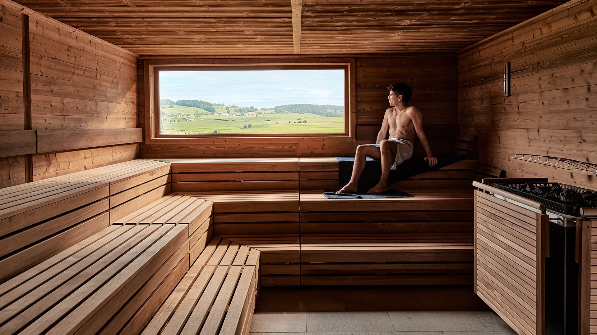 Mann sitzt auf Bank in Sauna mit Blick auf grüne Landschaft durch großes Fenster.