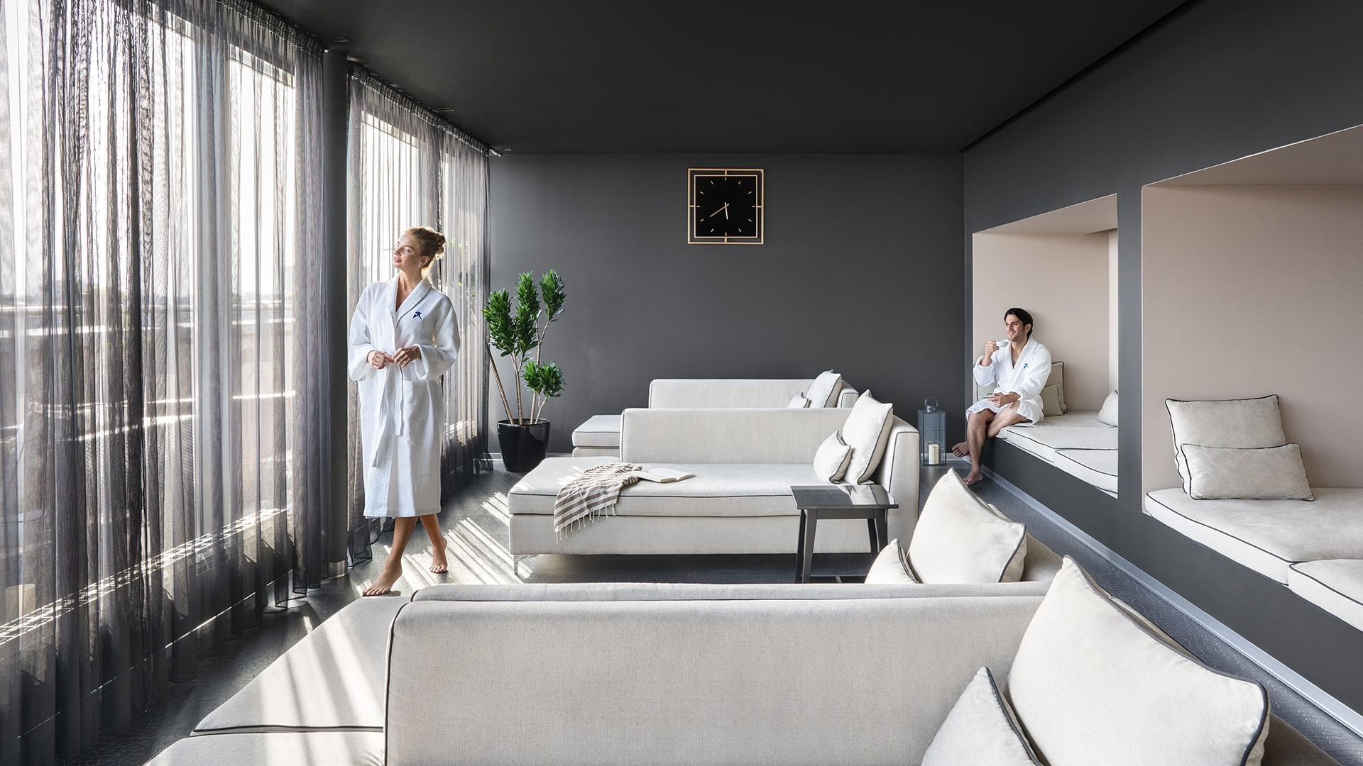 Frau im Bademantel in modernem Raum mit Sofas, Pflanzen und Uhr an der Wand.
