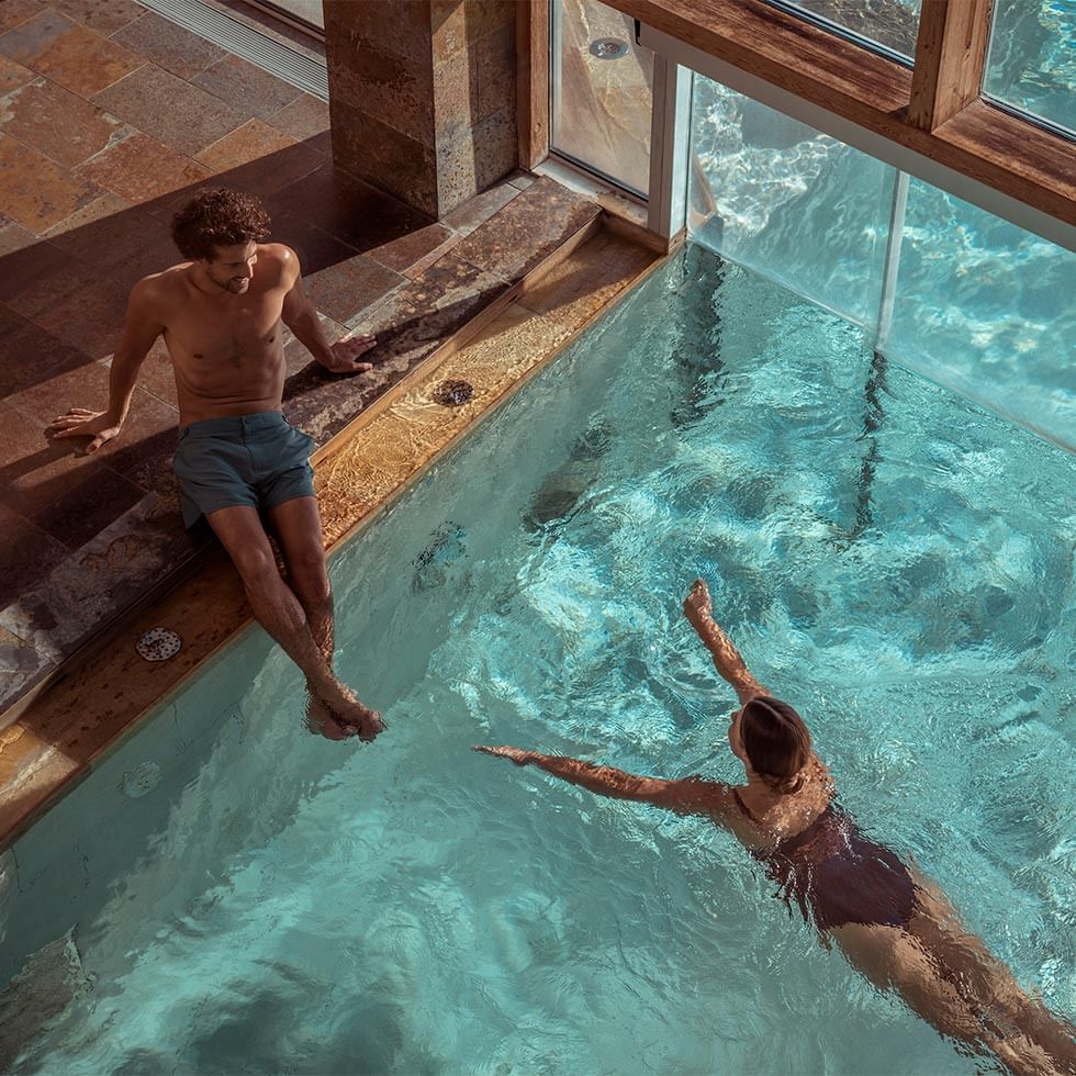 Mann und Frau in einem Hallenbad, er sitzt am Rand, sie schwimmt.