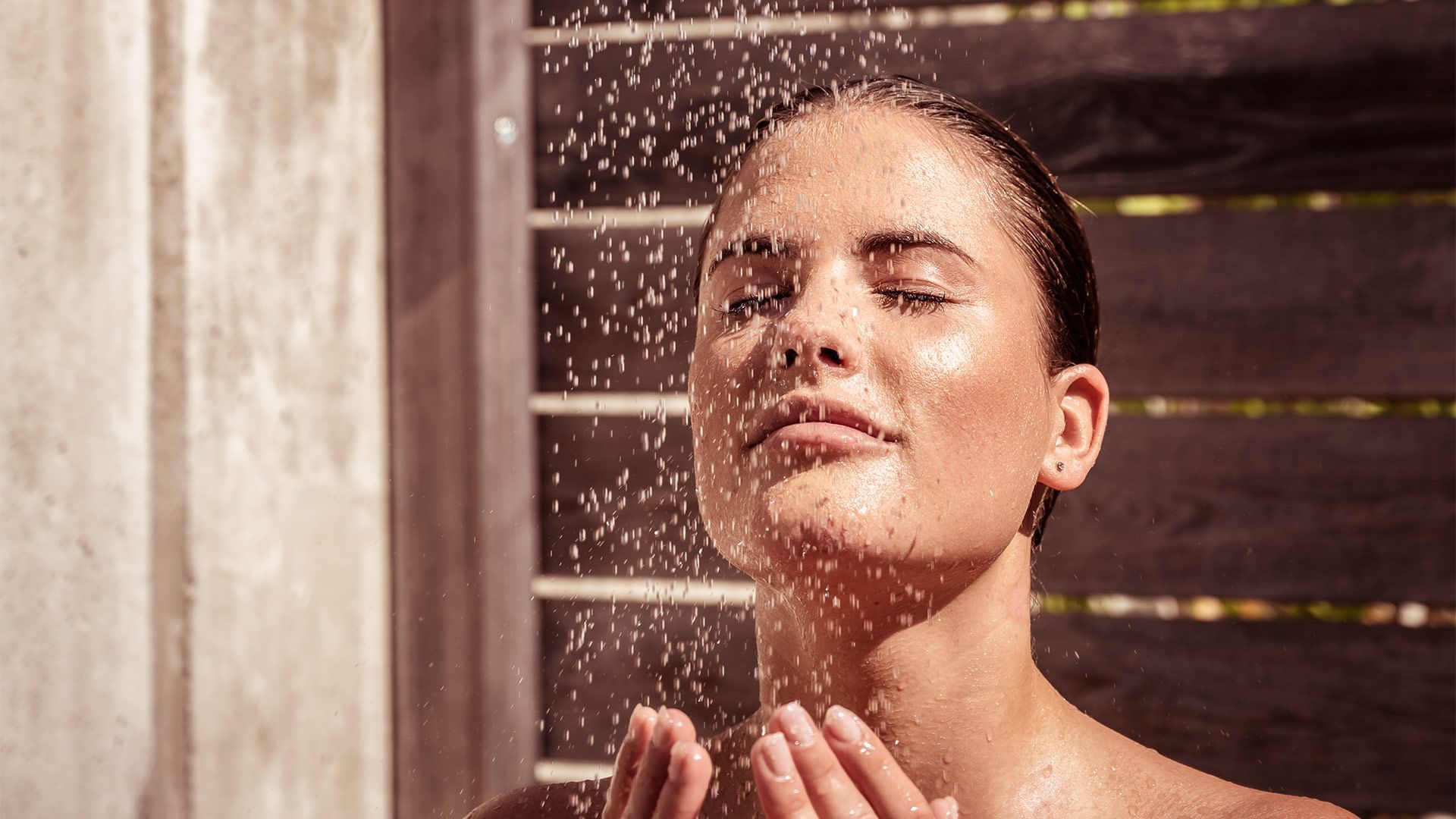 Eine Frau genießt eine Dusche mit Wassertropfen auf ihrem Gesicht und ihren Händen vor einer hölzernen Wand.