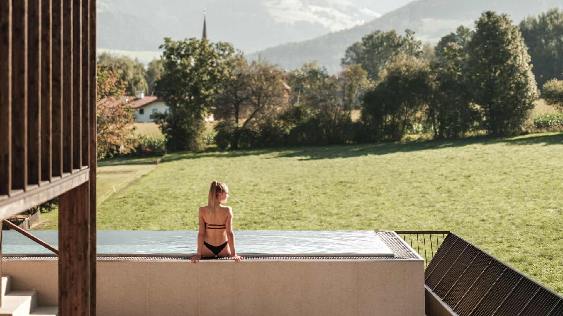 Frau in Bikini sitzt am Rand eines Schwimmbads und genießt die Aussicht auf grüne Felder und Berge.