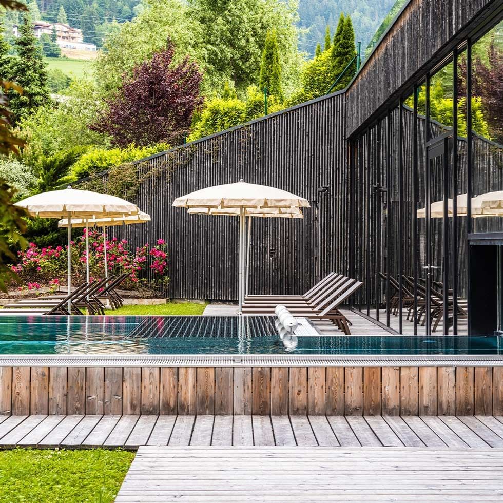 Modernes Schwimmbad mit Sonnenschirmen und Liegestühlen, umgeben von Bäumen und einer schwarzen Holzwand.