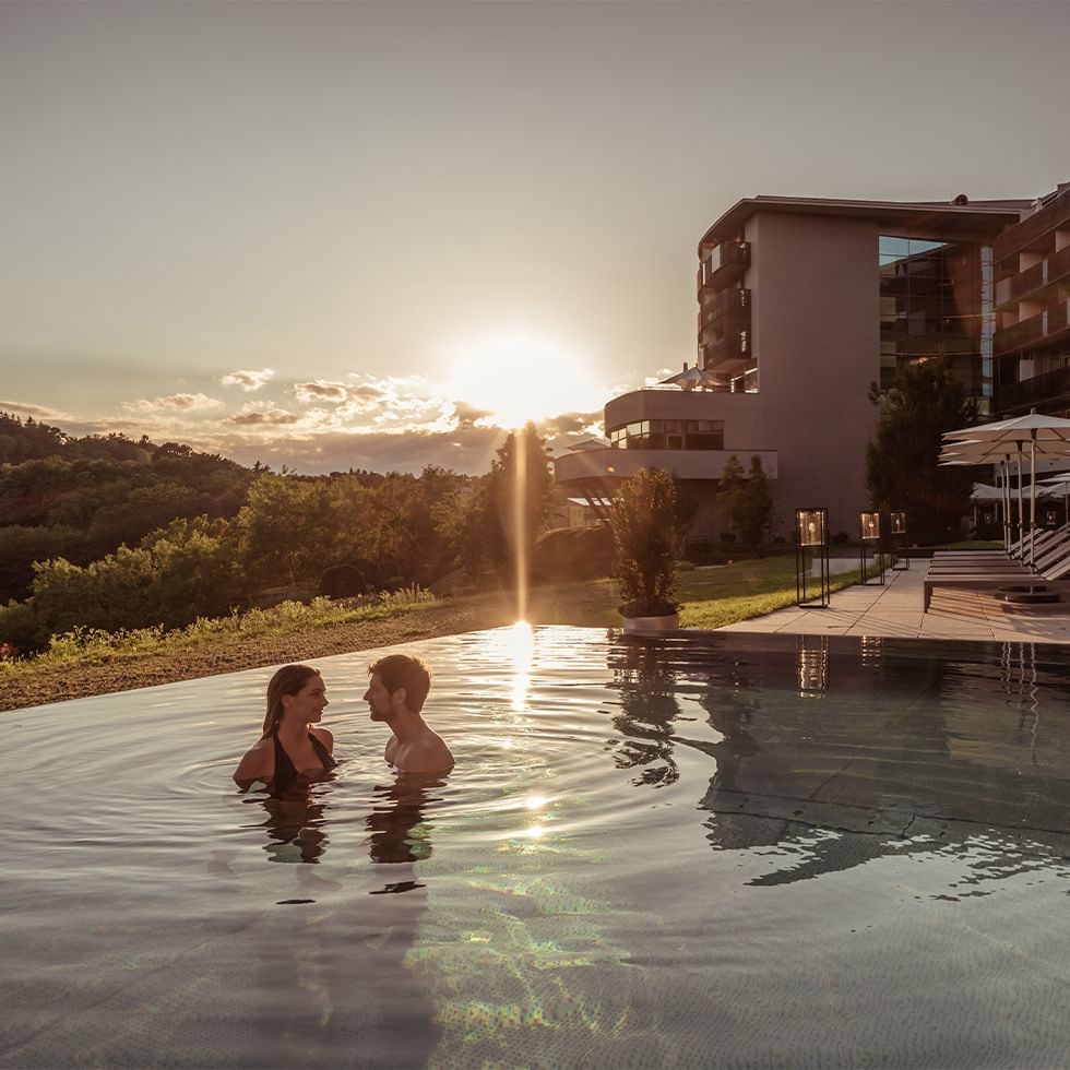 Zwei Personen genießen den Pool in einem Resort bei Sonnenuntergang, umgeben von Bäumen und Gebäuden.