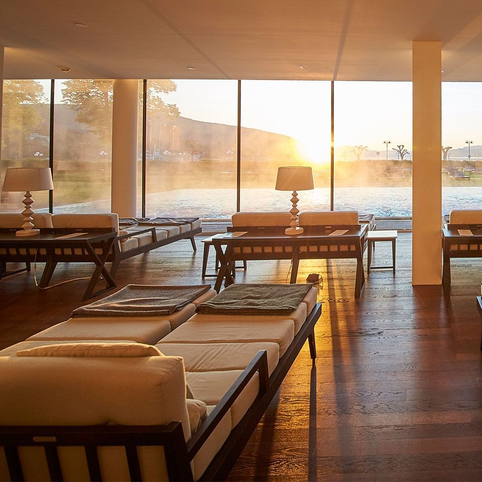 Loungebereich mit großen Fenstern, Couches, Tischen und Lampen, Blick auf Wasser und Sonnenuntergang.