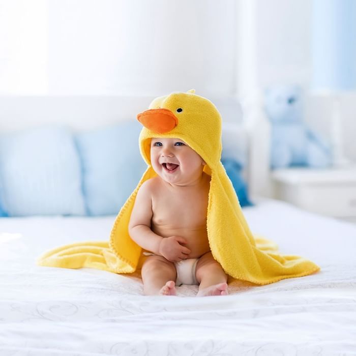 Ein Baby, das in eine gelbe Entenhandtuch gewickelt ist, sitzt lächelnd auf einem Bett.