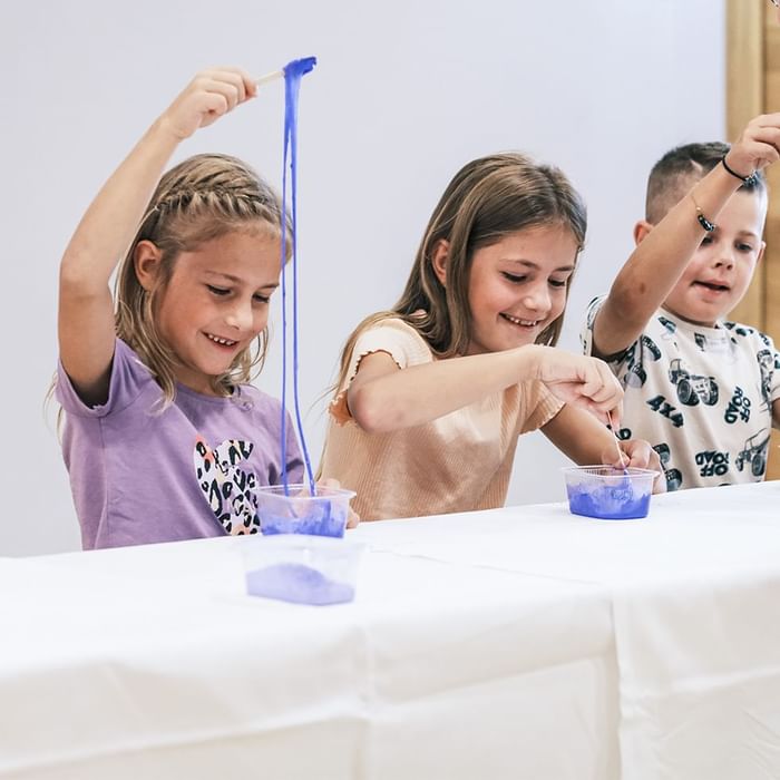 Drei Kinder spielen mit blauer Flüssigkeit in Behältern an einem Tisch.