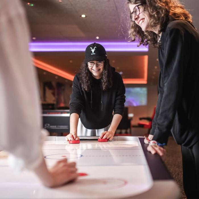 Drei Personen spielen Tisch-Airhockey in einem Spielzimmer, wobei eine Frau den Puck bewegt.