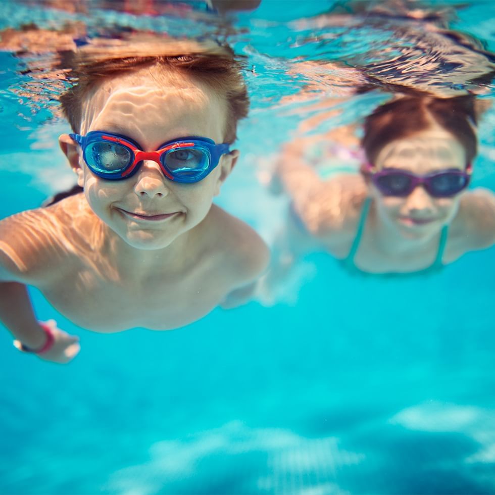 Zwei Kinder mit Schwimmbrillen, die unter Wasser im Schwimmbad lächeln.