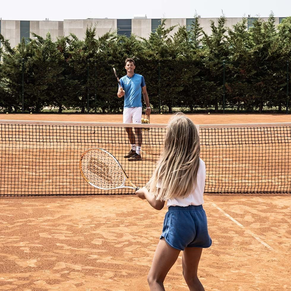 Mädchen mit Tennisschläger auf Sandplatz, Mann mit Schläger hinter Netz, Bäume und Gebäude im Hintergrund.