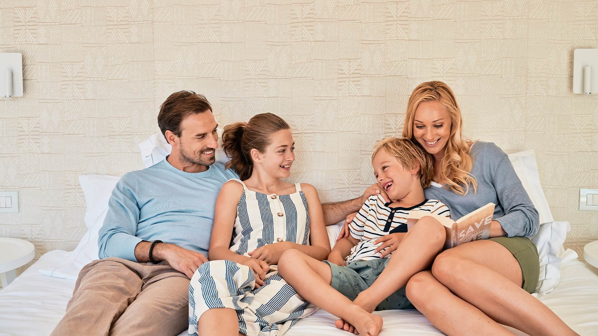 Familie auf Bett, Mutter liest Buch vor, Kinder lachen, Eltern lächeln, strukturiertes beiges Kopfteil.