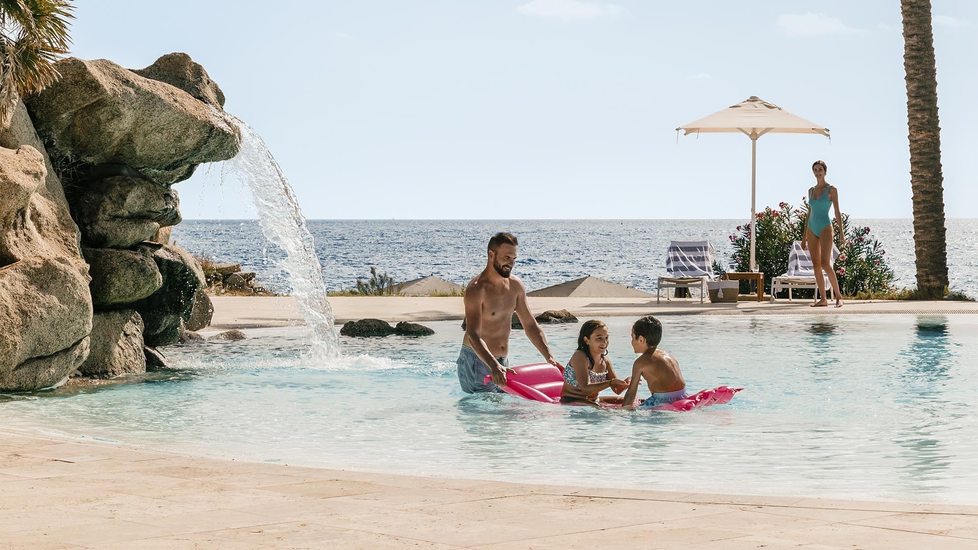 Familie genießt den Pool mit Wasserfall neben dem Meer, während eine Frau im Hintergrund spaziert.