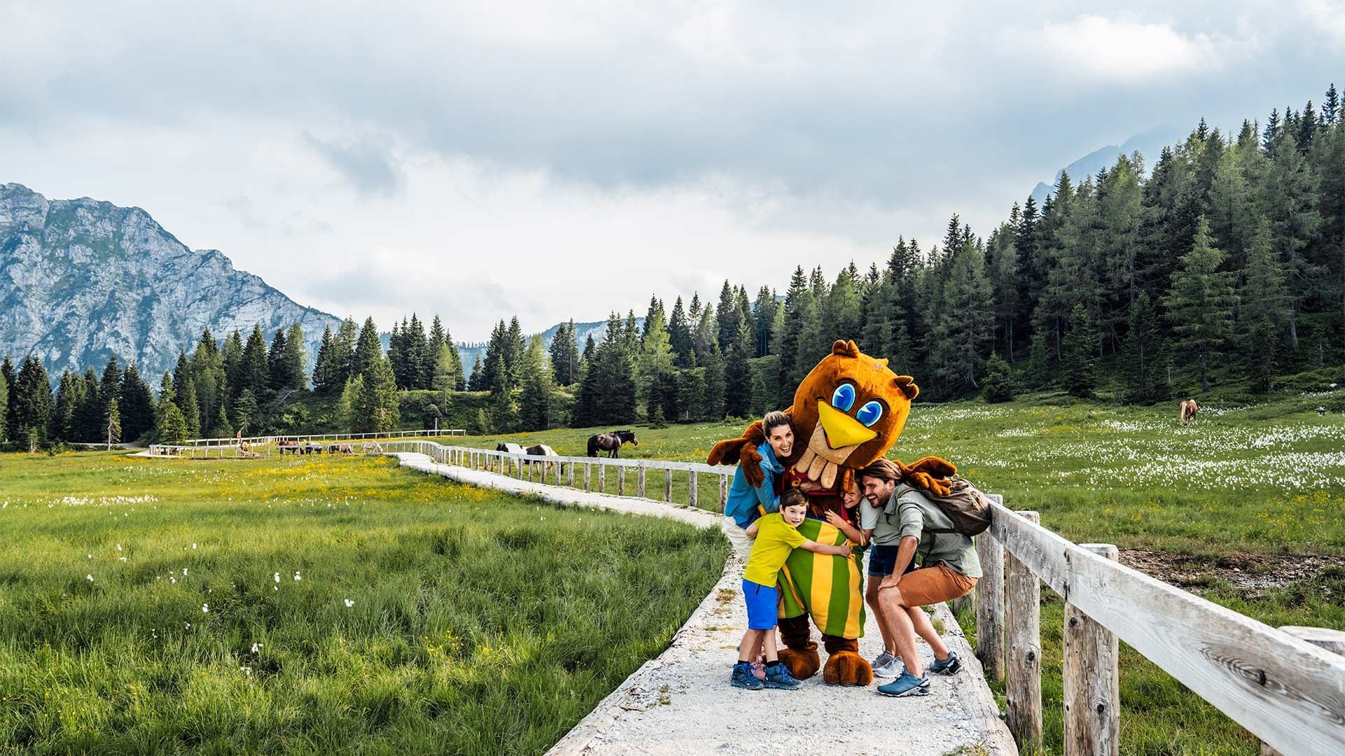 Familie und Maskottchen umarmen sich auf einem Pfad im Grünen vor einer Waldkulisse.