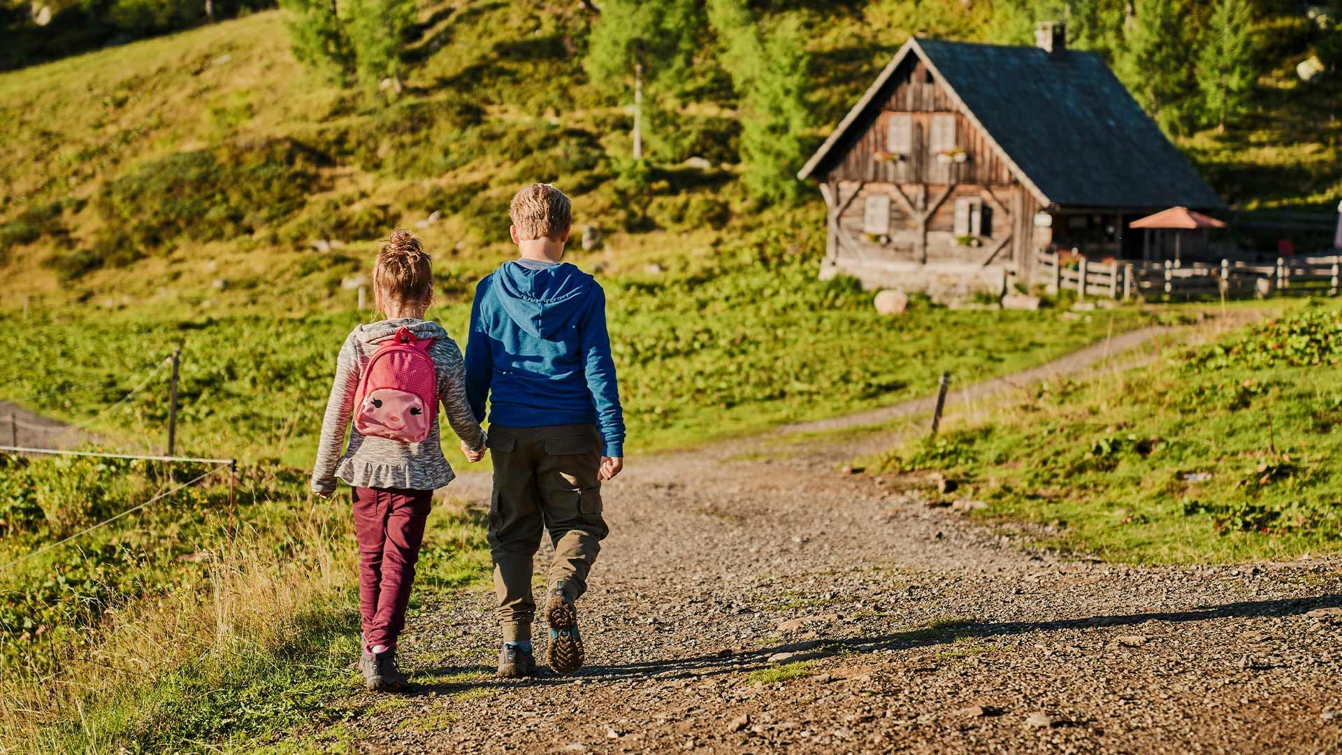 Zwei Kinder gehen Hand in Hand auf einem Pfad neben einem hölzernen Haus.