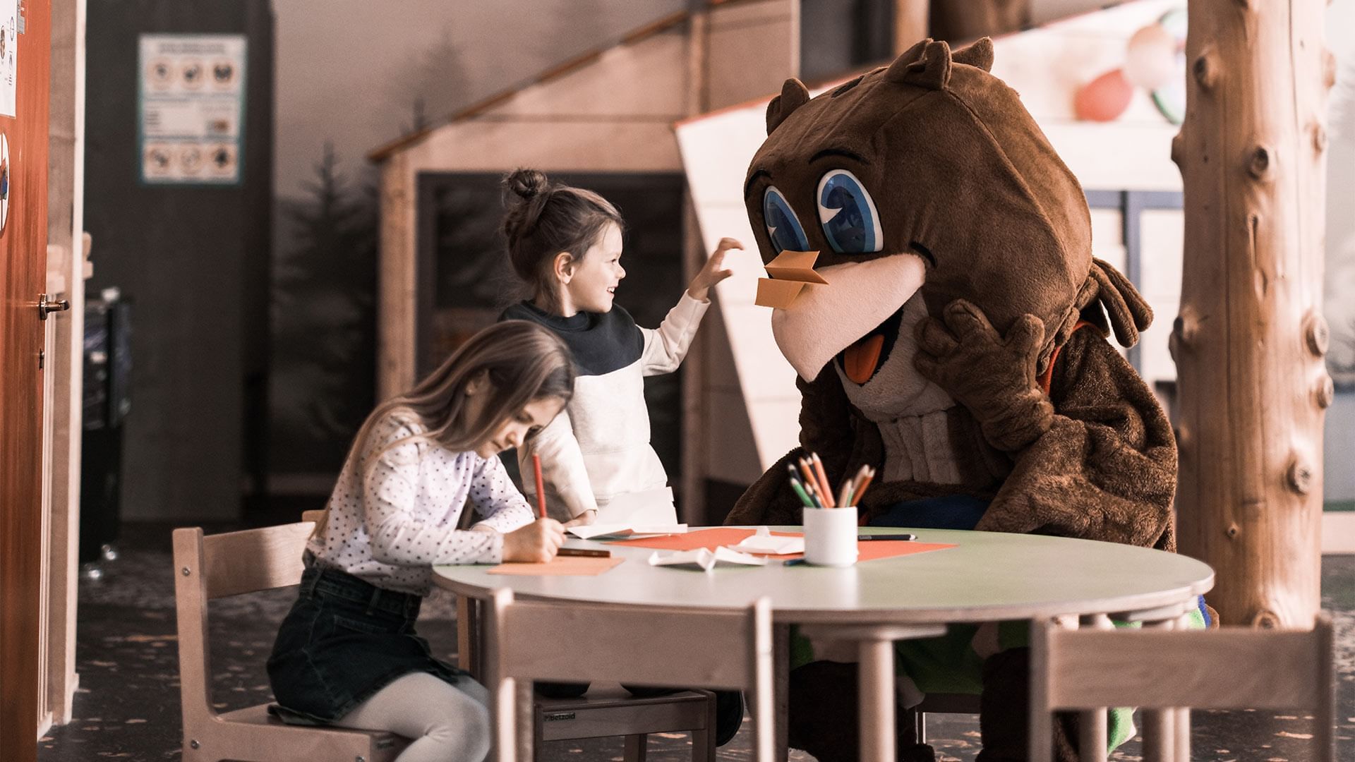 Zwei Mädchen schreiben mit Stiften an einem Tisch, während ein Maskottchen daneben sitzt.
